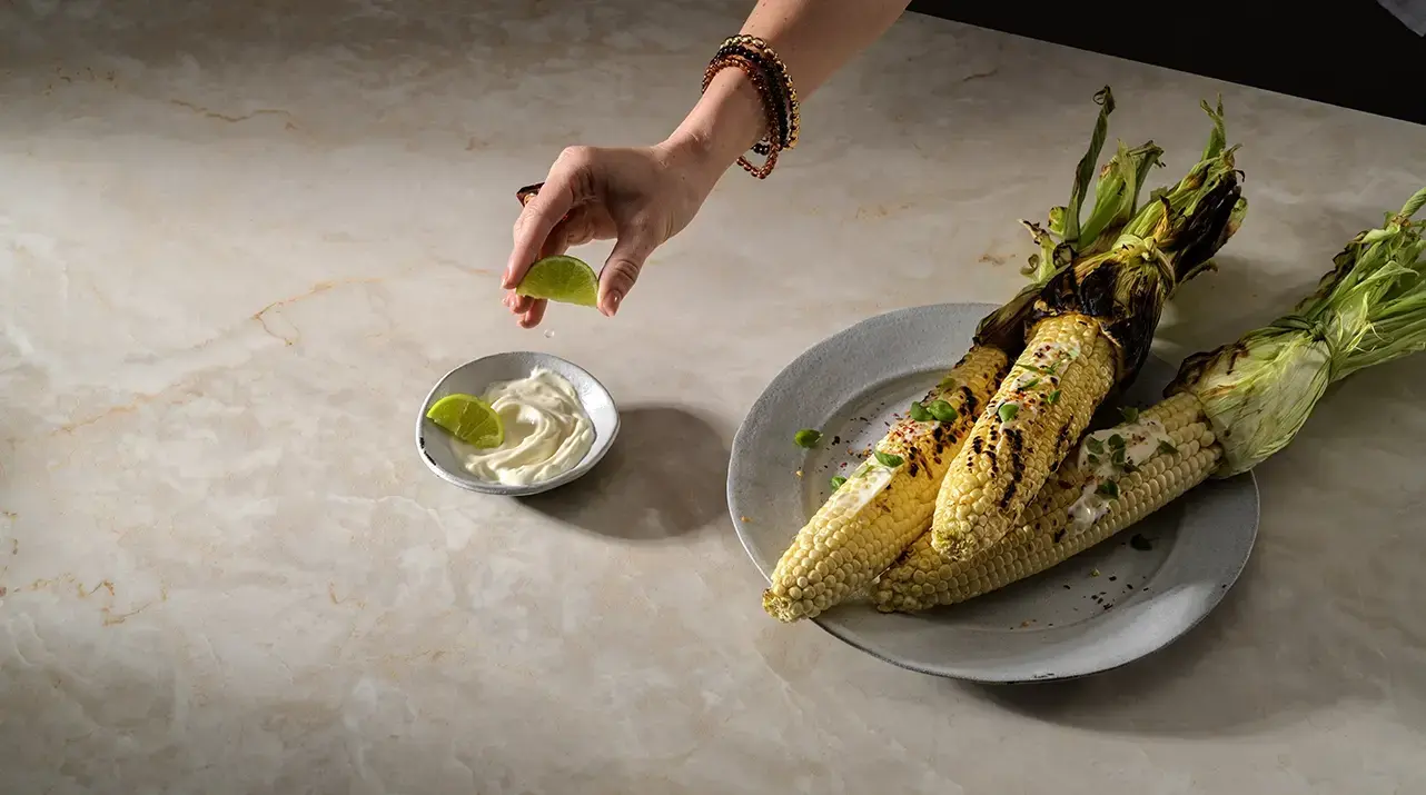Caesarstone 583 Crestone porcelain countertop surface featuring a soft alabaster base with delicate, translucent veining, presented in a lifestyle culinary setting with a plate of grilled corn.