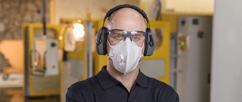 Fabricator wearing protective gear including a respirator mask, safety glasses, and earmuffs, standing confidently in a Caesarstone workshop.