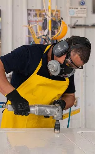 Worker wearing protective gear and yellow apron uses power tool on a white surface.