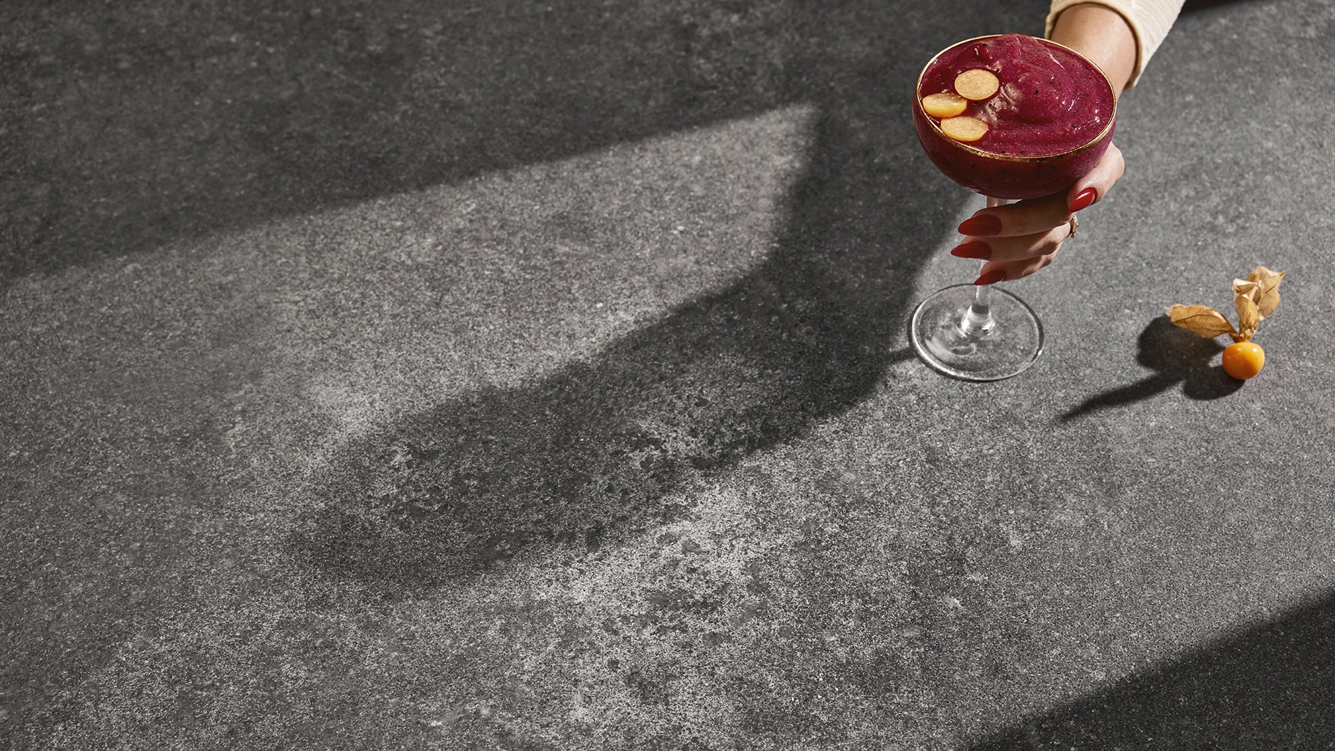 Hand with a dark berry cocktail resting on Caesarstone 4033 Rugged Concrete countertop, highlighting its rugged matte texture and natural concrete look.